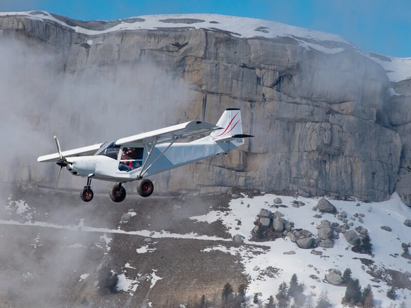 Avion ULM blanc volant dans un paysage montagneux avec des falaises et de la neige sous un ciel partiellement nuageux.