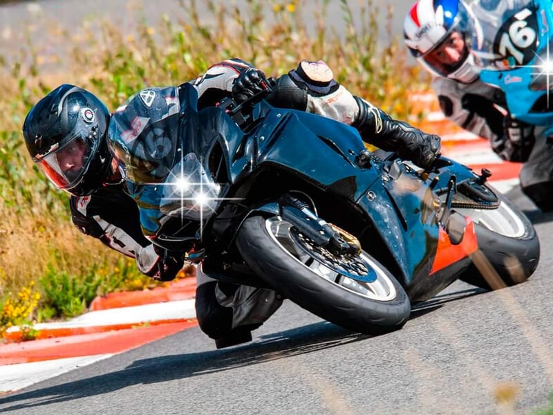 Compétition moto sur circuit, pilote en combinaison noire au guidon d'une moto noire inclinée à l'extrême, suivi de près par un concurrent en moto bleue et blanche