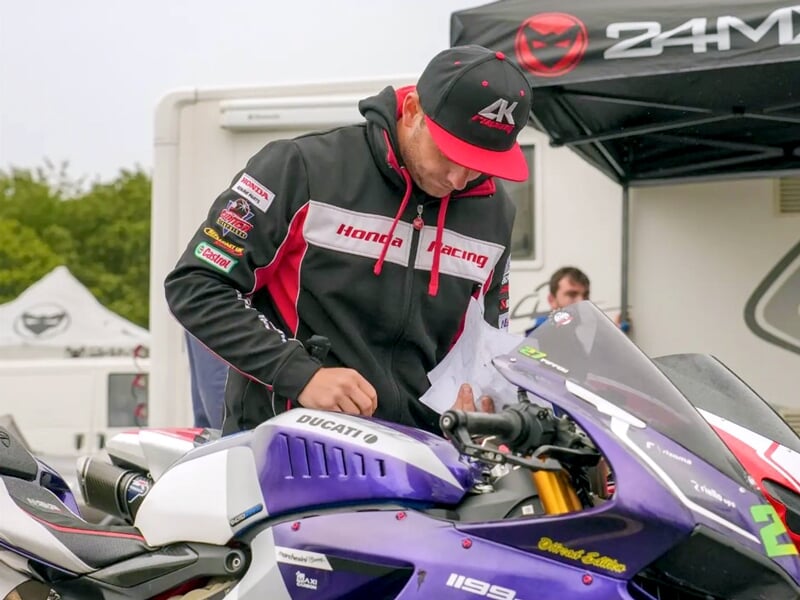 Pilote en tenue Honda Racing consultant des documents devant une moto Ducati 1199 Panigale violette et blanche, dans un paddock de course avec des tentes et camions en arrière-plan