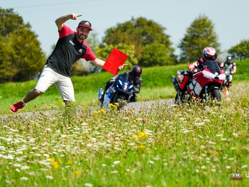 Encadrement de stage moto, un animateur en tenue sportive brandit un drapeau rouge au bord de la piste tandis que des motards en pleine accélération approchent sur un circuit entouré de verdure.