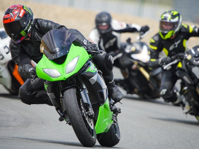 Course moto sur circuit, pilote en combinaison noire au guidon d'une Kawasaki verte inclinée dans un virage serré, suivi de plusieurs concurrents en pleine trajectoire.
