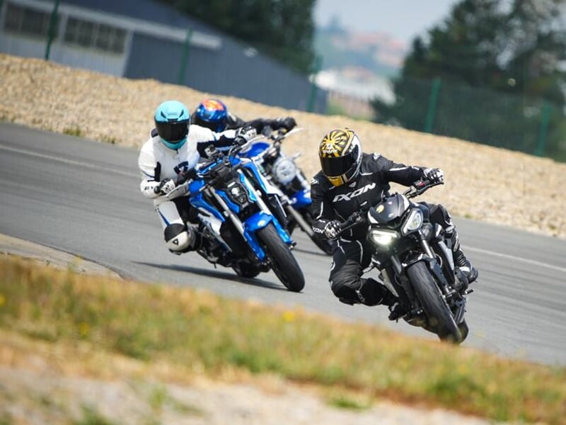 Entraînement sur circuit, motard en combinaison noire et casque jaune au guidon d'une moto noire suivi de pilotes en roadster bleus et blanc, en plein virage.
