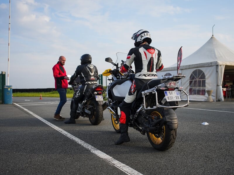 Préparation avant roulage, deux motards en combinaison sur des motos routières attendent les instructions d'un encadrant en veste rouge près d'une tente sur un paddock.