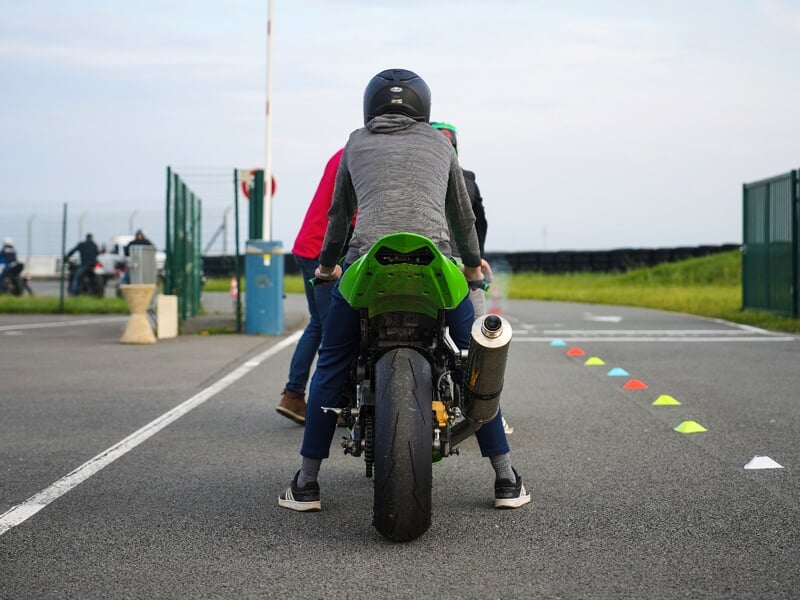 Départ d'entraînement moto, pilote vu de dos sur une sportive verte prêt à s'élancer, aligné avec des cônes de signalisation, tandis que d'autres roulent en arrière-plan.