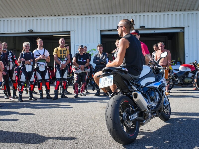 Session de briefing moto, un instructeur assis sur une BMW S1000RR face à un groupe de participants en tenue de piste devant un garage du circuit.