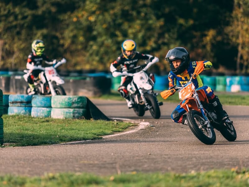 Course de mini-motos sur circuit, un enfant en combinaison jaune et bleue mène la course, suivi de deux concurrents en tenue noire et blanche.