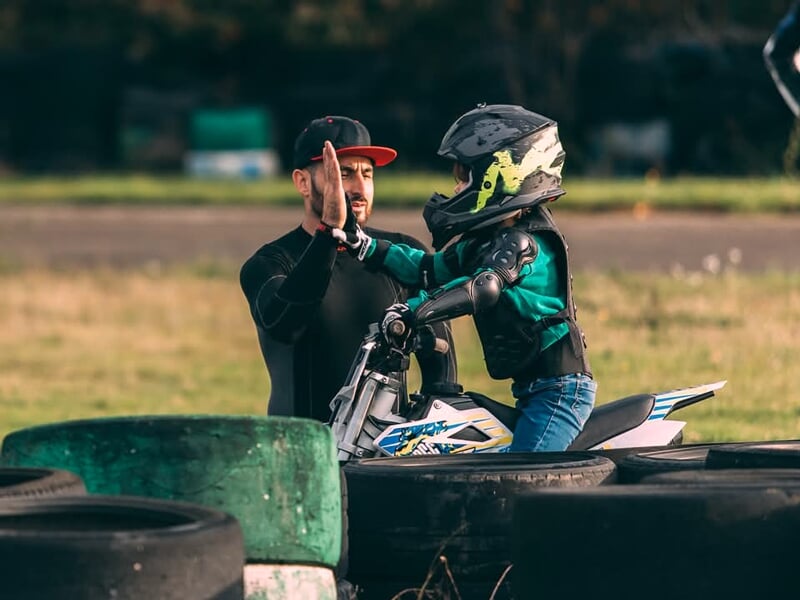 Séance d’apprentissage moto, un enfant en tenue noire et verte sur une mini-moto blanche reçoit un encouragement par un high-five d’un instructeur en noir près des pneus de sécurité.
