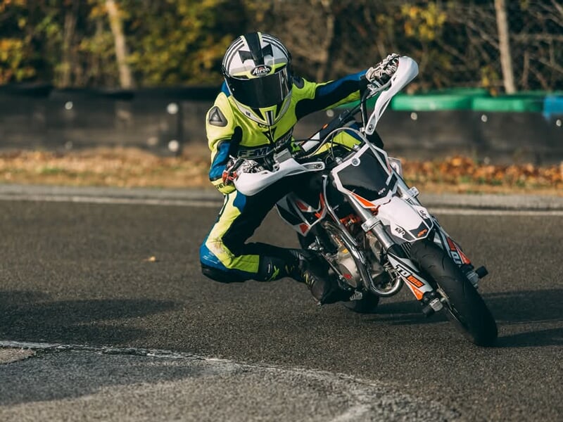 Motard en combinaison jaune et bleue effectuant une inclinaison extrême sur une mini-moto blanche, maîtrisant son virage avec précision sur circuit.
