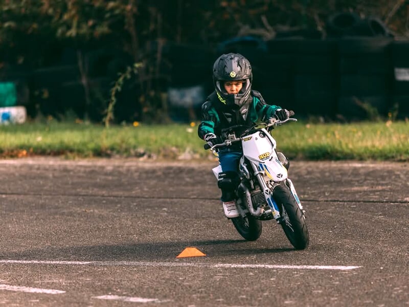 Enfant en combinaison noire et verte pilotant une mini-moto blanche entre des cônes de signalisation sur un circuit d’apprentissage, concentré sur sa trajectoire.