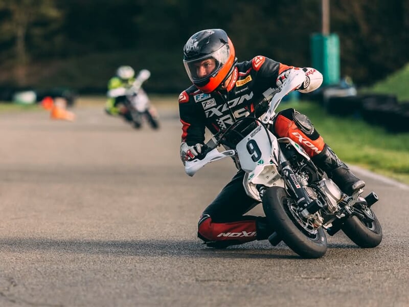 Pilote en combinaison rouge et noire penché au ras du sol sur une mini-moto blanche, en pleine prise d’angle sur circuit, avec un autre concurrent en arrière-plan.