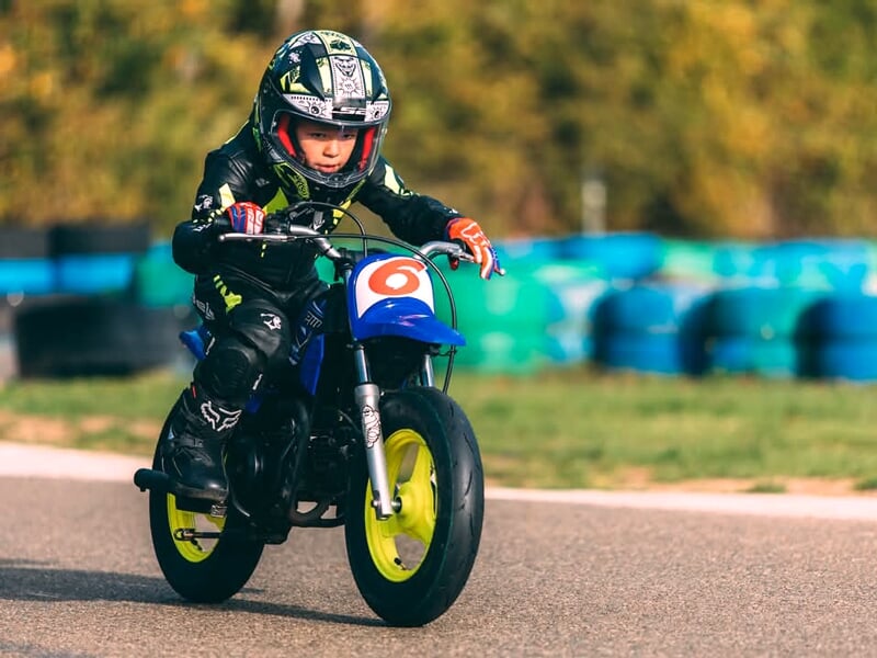 Enfant en équipement moto noir et vert concentré au guidon d’une mini-moto bleue aux roues jaunes, roulant sur un circuit entouré de pneus colorés.