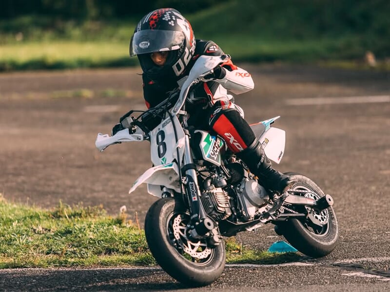 Jeune pilote en combinaison noire et rouge maîtrisant une mini-moto blanche lors d’un virage serré sur piste, incliné pour optimiser sa trajectoire.