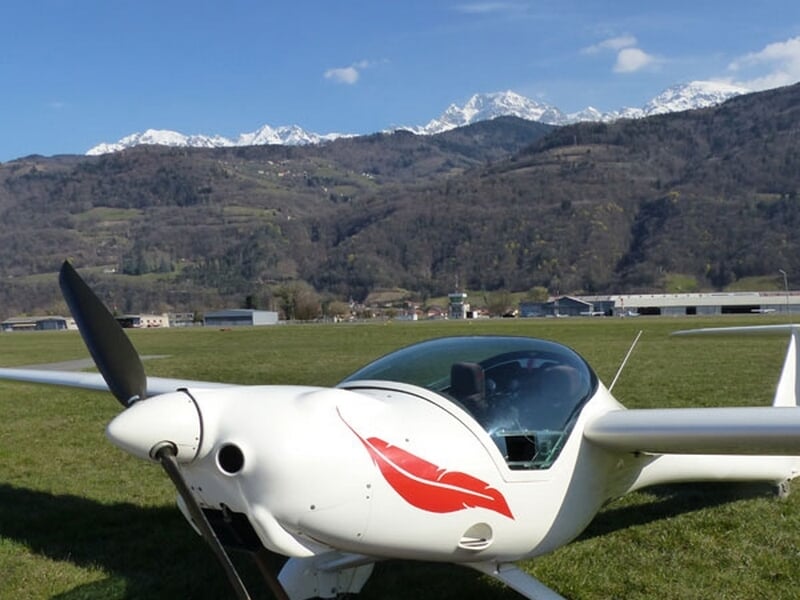 Planeur blanc au sol avec un dessin rouge, montagnes enneigées en arrière-plan sous un ciel bleu clair.