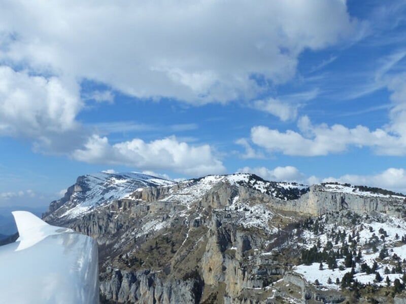 Vue en vol d'un planeur au-dessus d'une montagne rocheuse partiellement enneigée sous un ciel nuageux.