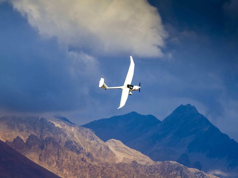 Planeur blanc en vol avec hélice visible, survolant des montagnes avec un ciel nuageux et sombre.