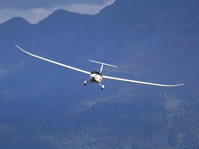 Planeur vu de face en vol, ailes déployées, avec montagnes en arrière-plan sous un ciel bleu clair.