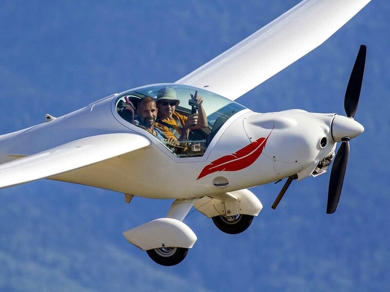 Deux personnes dans un planeur blanc en vol, l'une fait un signe de la main, ciel bleu et montagnes en arrière-plan.