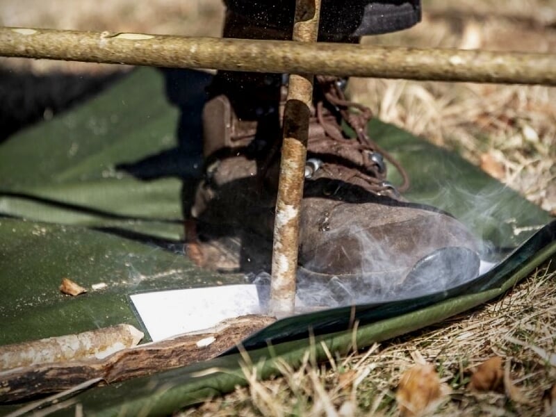 Personne allumant un feu de camp avec un bâton et un support, fumée visible au sol en forêt.