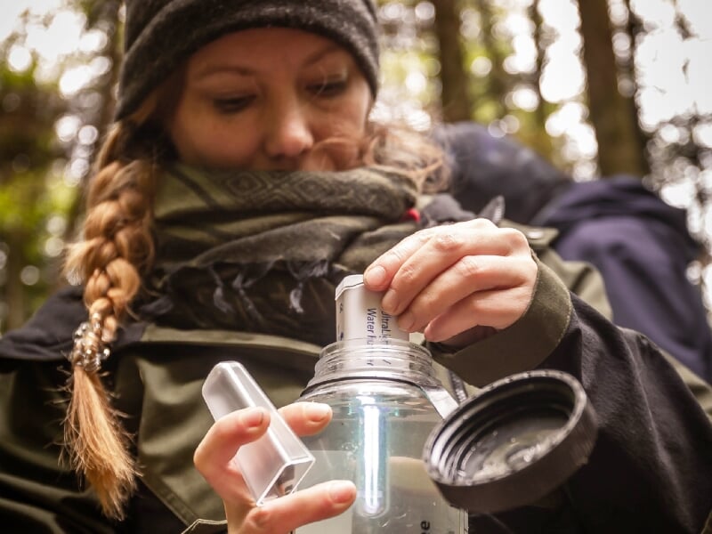 Femme en tenue de randonnée versant de l'eau dans une gourde transparente, hydratation en nature.