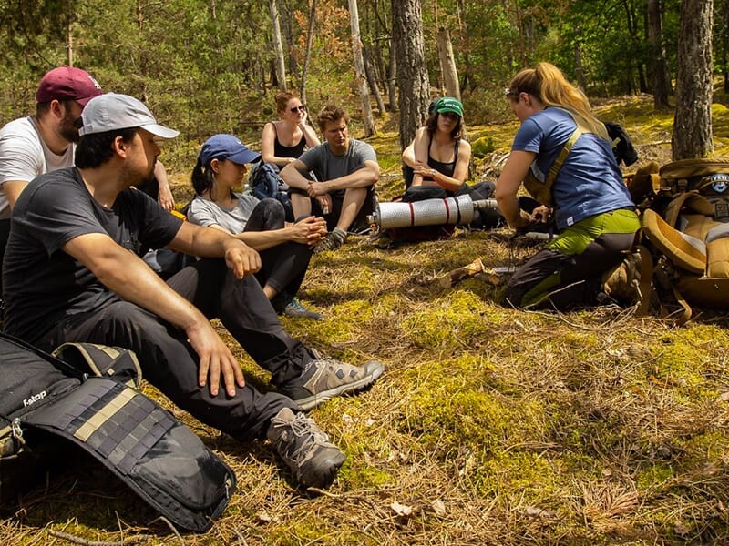 Groupe de personnes assises en cercle dans une forêt, participant à un stage de survie près de Vesoul, avec équipement et sac à dos.