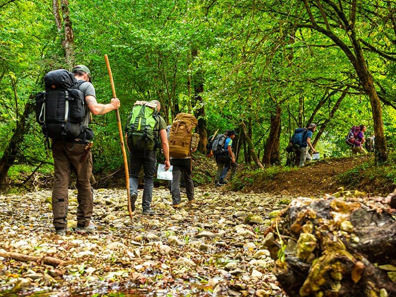 Groupe de randonneurs avec de grands sacs à dos traversant un lit de rivière rocheux en pleine nature.