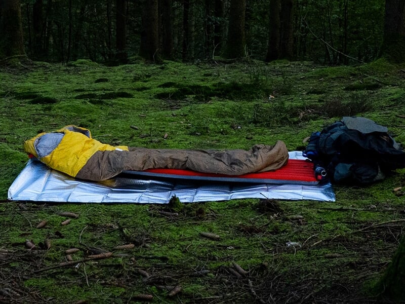Tapis de sol en pleine forêt avec un sac de couchage déplié, prêt pour le bivouac.