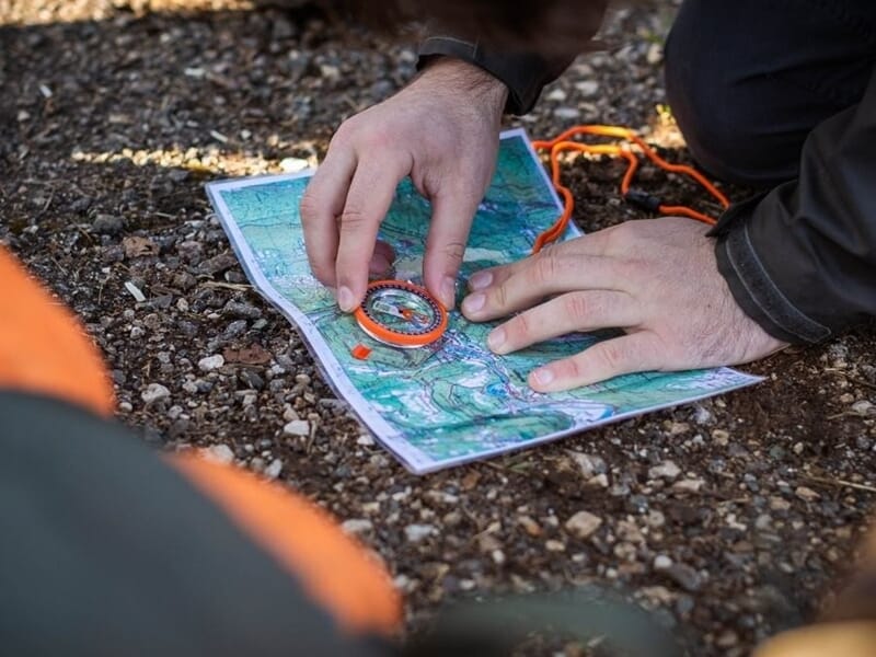 Main utilisant une boussole posée sur une carte topographique lors d'un stage de survie dans les Vosges du Nord.