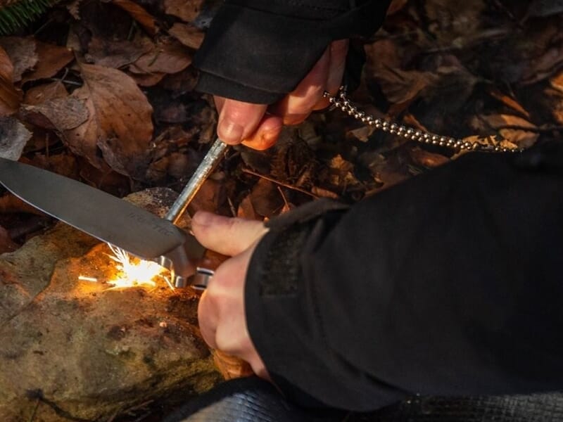 Personne en tenue noire allumant un feu avec un couteau et un briquet sur un sol couvert de feuilles mortes en forêt