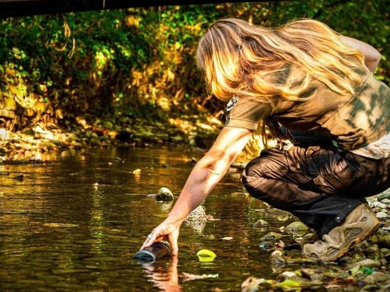 Personne accroupie au bord d'un ruisseau en forêt, touchant l'eau avec la main, lumière naturelle
