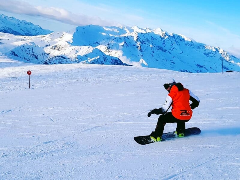 Snowboarder en tenue rouge et noire glissant sur une piste enneigée avec montagnes en arrière-plan.