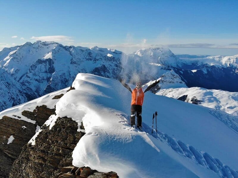 Skieur en veste orange levant les bras au sommet d'une montagne enneigée avec vue panoramique.