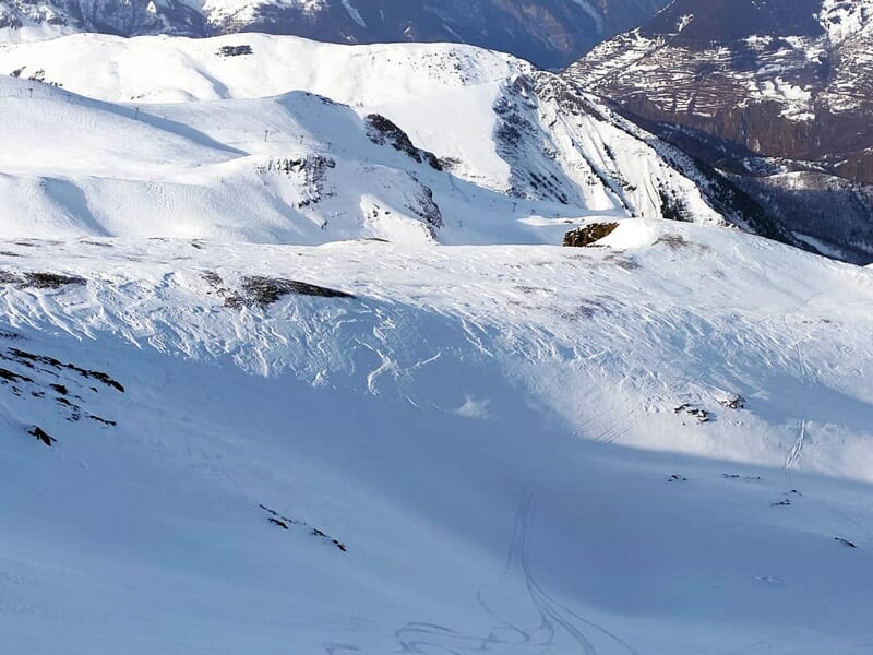 Paysage de montagne enneigée avec traces de ski et vallées visibles sous un ciel clair.