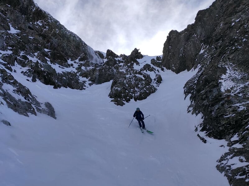 Skieur descendant une pente étroite et enneigée entre des falaises rocheuses dans les Alpes.