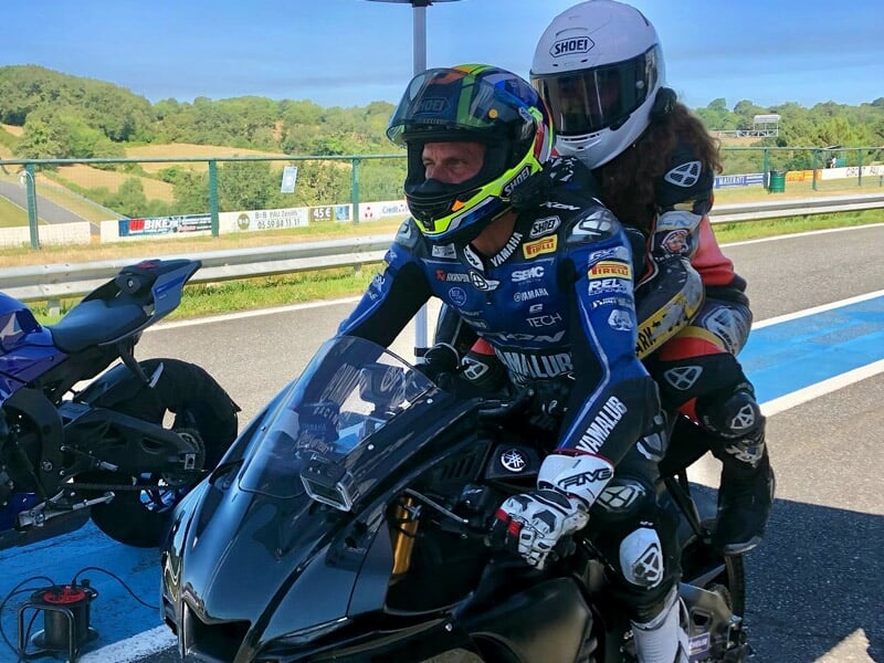 Deux motards en tenue de course préparant une moto noire dans une allée de stand, un avec un casque intégral blanc