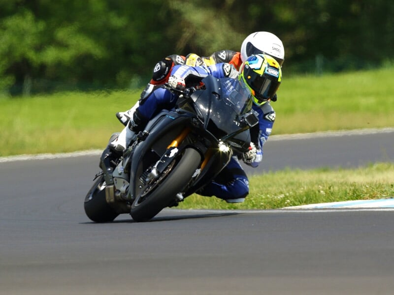 Pilote de moto en combinaison de course blanche et noire prenant un virage serré sur une piste de course avec des gradins colorés en arrière-plan