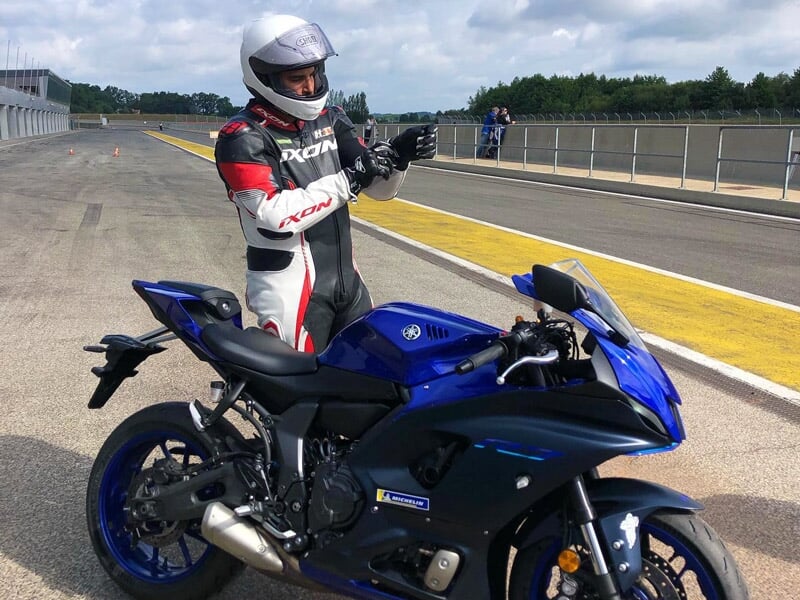 Un pilote en combinaison de course blanche, rouge et noire se préparant à monter sur sa moto de sport Yamaha bleue sur la ligne de départ d'un circuit de course