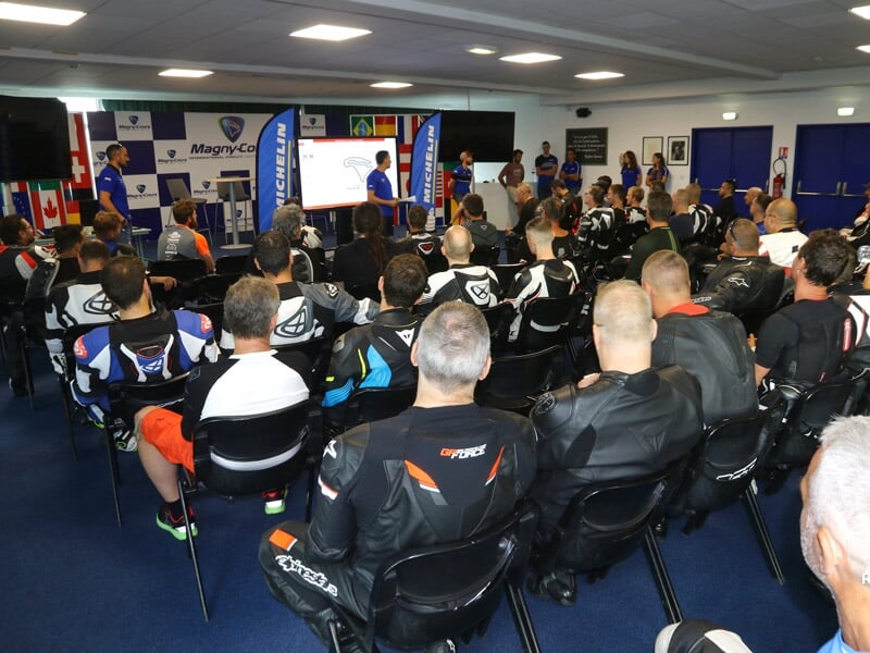 Salle de briefing avec des pilotes de moto en combinaisons de course écoutant une présentation avant la course