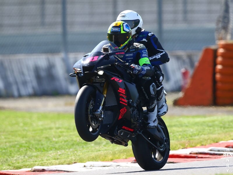 Pilote de moto en tenue de course bleue et noire, casque blanc, effectuant un wheelie sur une piste de course