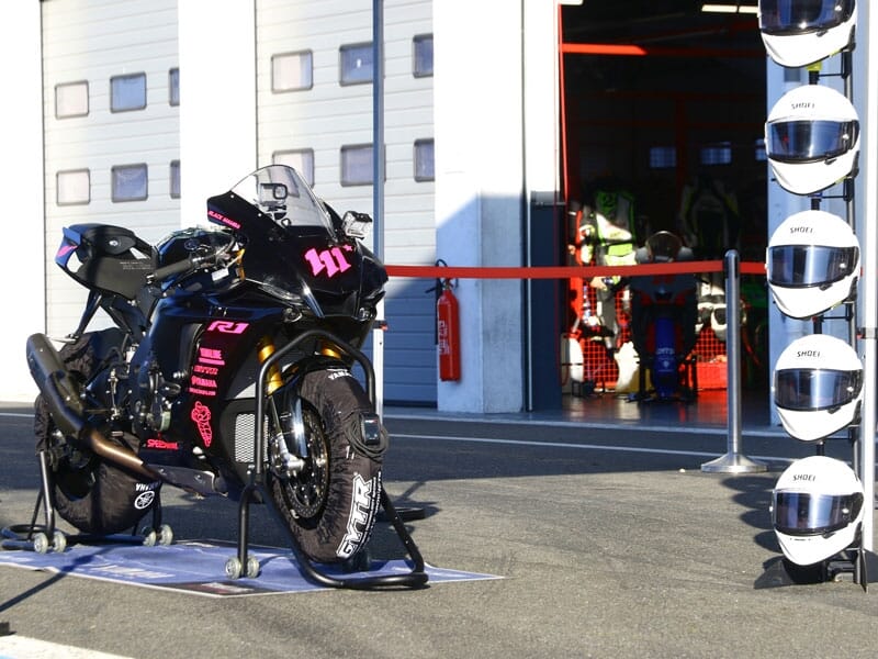 Moto de course noire stationnée devant un garage avec une rangée de casques sur un support à côté