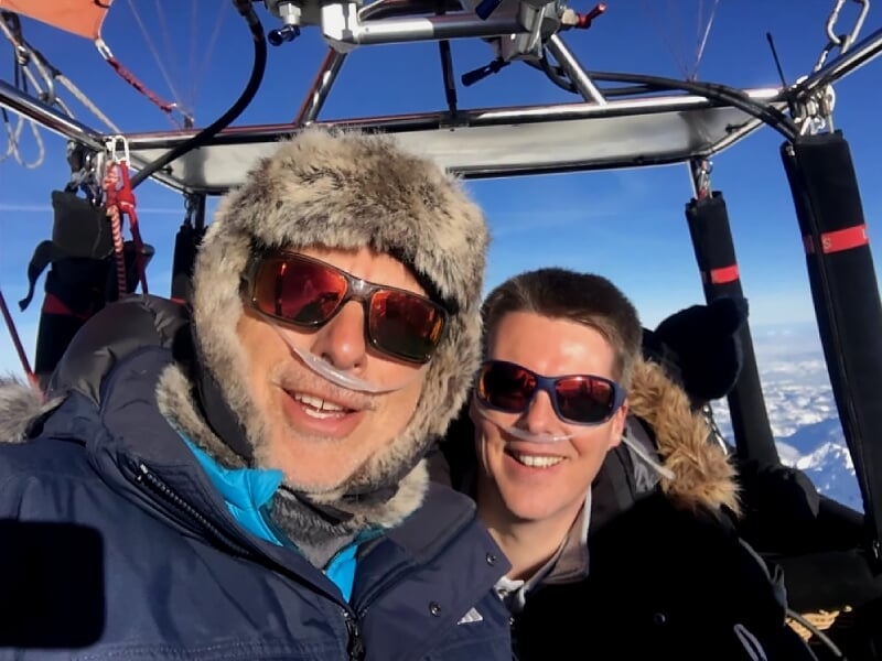 Deux personnes souriantes en tenue d'hiver dans la nacelle d'une montgolfière en plein vol.
