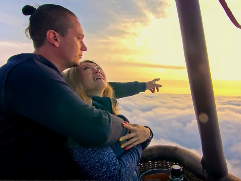 Couple dans une montgolfière au coucher du soleil, la femme regarde vers l'horizon en souriant.