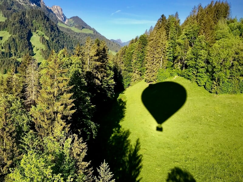 Ombre d'une montgolfière projetée sur une prairie verte entourée d'arbres en montagne.