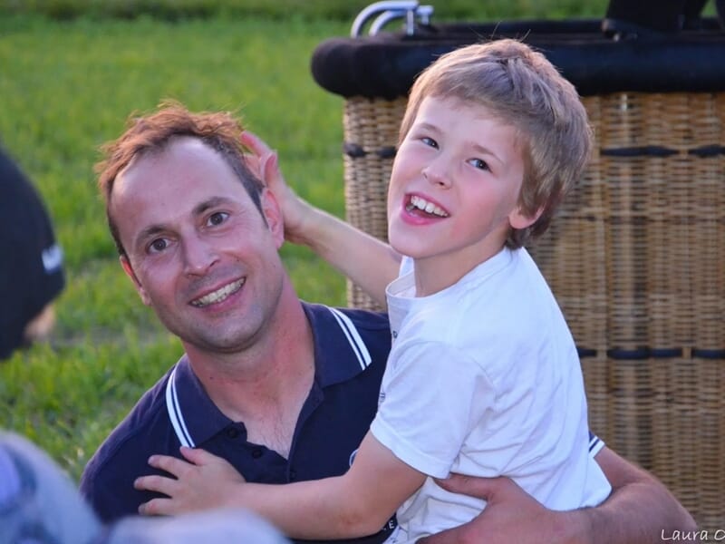 Un homme souriant tient un garçon joyeux dans ses bras devant une montgolfière en préparation dans un champ vert.