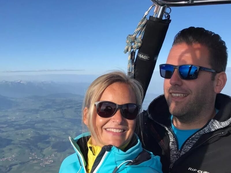 Un couple souriant porte des lunettes de soleil dans une nacelle de montgolfière avec un paysage de montagnes en arrière-plan.