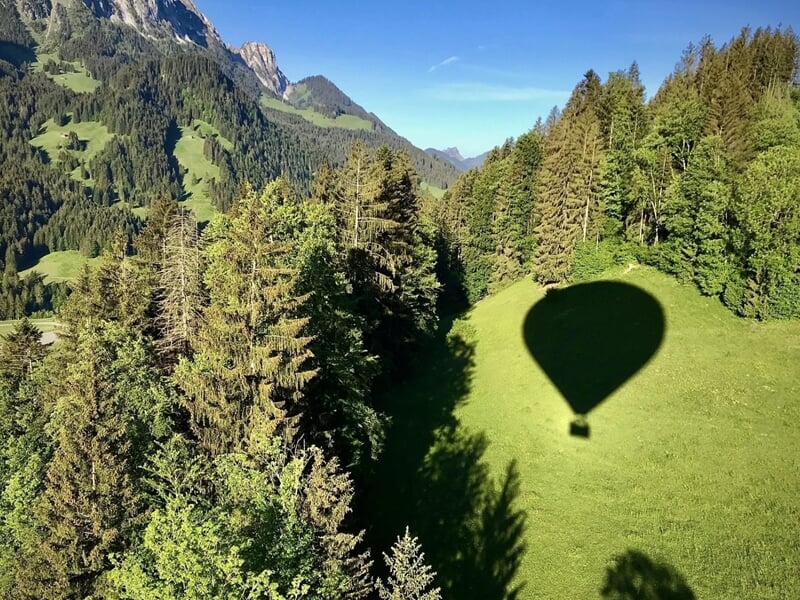 Ombre d'une montgolfière projetée sur une prairie entourée de forêts et montagnes lors d'un vol en montgolfière dans le Chablais.