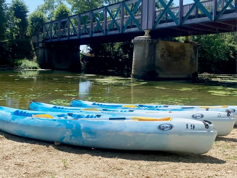 Trois kayaks bleus posés sur le bord d'une rivière calme sous un pont en métal entouré d'arbres verts.