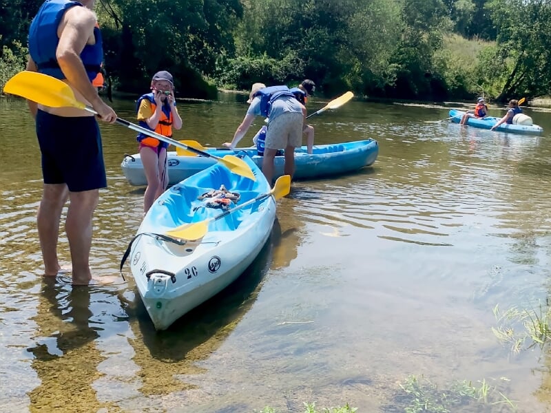 Des personnes équipées de gilets de sauvetage embarquent dans des kayaks bleus sur une rivière paisible en été.