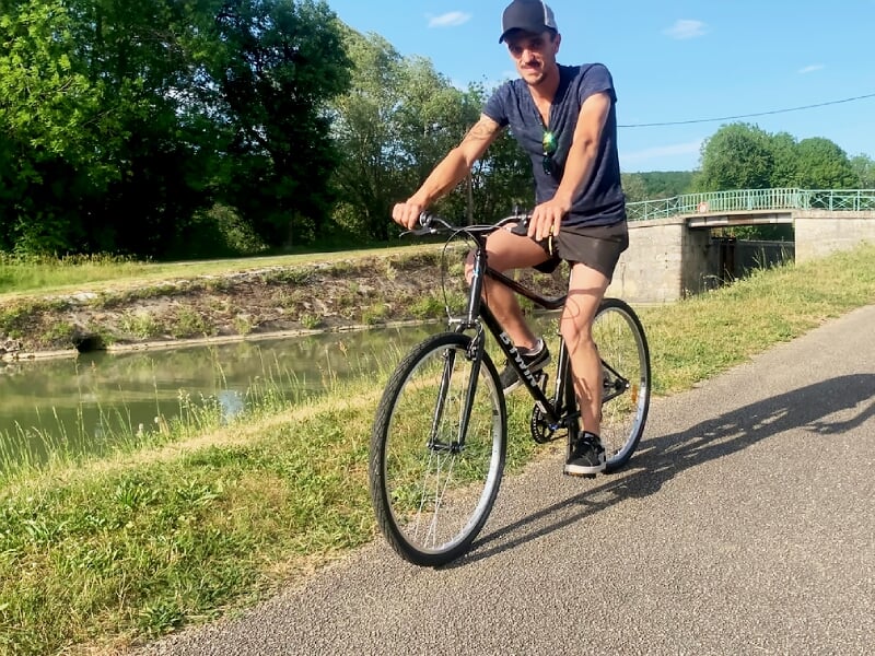 Un homme en tenue sportive roule à vélo sur une piste asphaltée le long d'un canal bordé d'herbe et d'arbres.