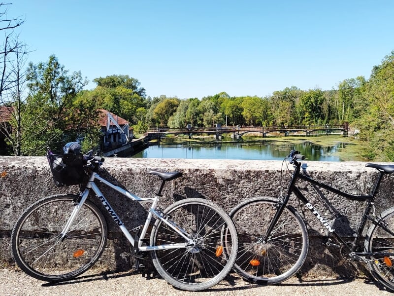 Deux vélos stationnés sur un pont en pierre avec vue sur un plan d'eau et des arbres sous un ciel bleu clair.