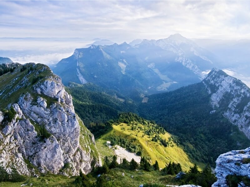 Vue aérienne d'un paysage montagneux verdoyant avec des sommets rocheux sous un ciel partiellement nuageux.